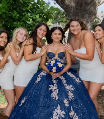 Quinceañera group portrait in a navy blue gown with gold lace, surrounded by friends in matching white dresses