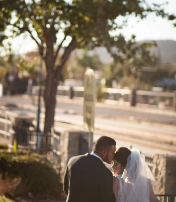 Wedding couple embracing near railing — groom kissing bride's forehead at golden hour