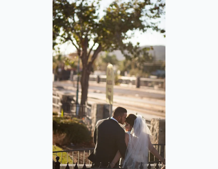 Wedding couple embracing near railing — groom kissing bride's forehead at golden hour