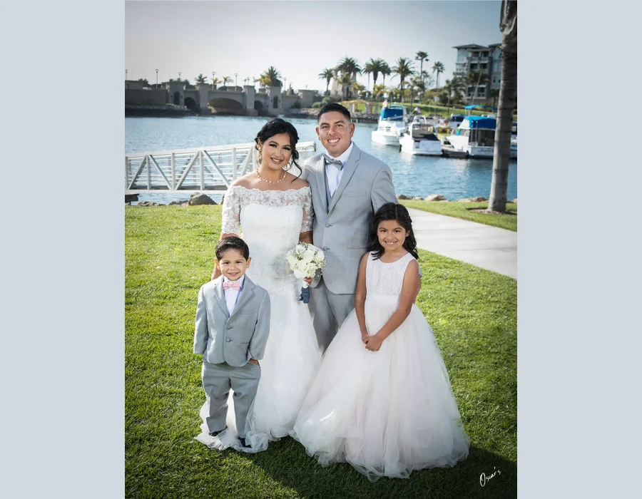 Wedding family portrait by the marina — bride, groom and two young children (ring bearer and flower girl) posing by a waterfront with boats and palm trees