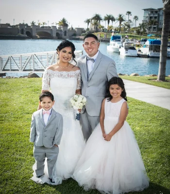 Wedding family portrait by the marina — bride, groom and two young children (ring bearer and flower girl) posing by a waterfront with boats and palm trees