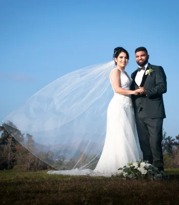 Wedding portrait with flowing veil — bride and groom holding hands outdoors, veil caught by the wind against a blue sky
