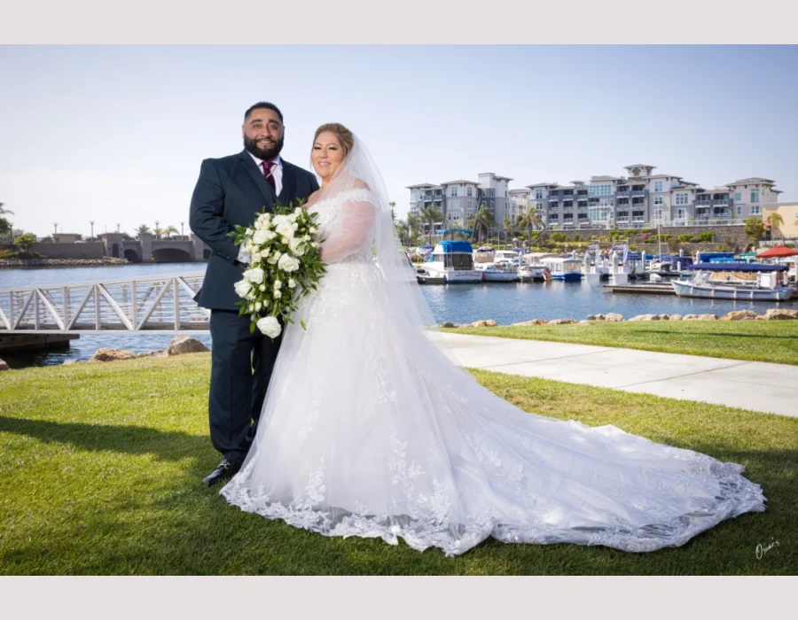 Wedding couple portrait by the harbor — bride in lace off-shoulder gown holding a white rose bouquet, groom in navy suit, marina in background
