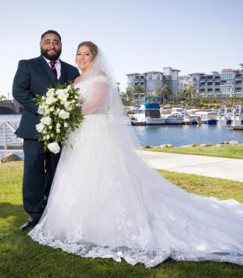 Wedding couple portrait by the harbor — bride in lace off-shoulder gown holding a white rose bouquet, groom in navy suit, marina in background