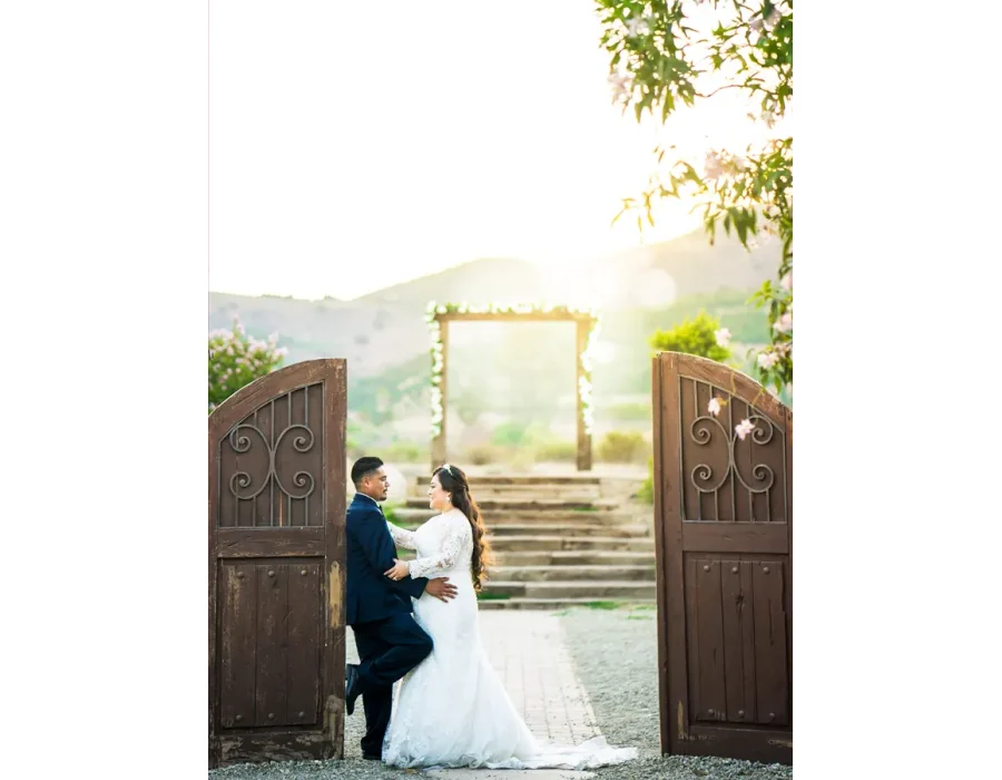 Wedding couple at a rustic wooden gate — bride and groom embracing at a wooden garden entrance with string lights beyond