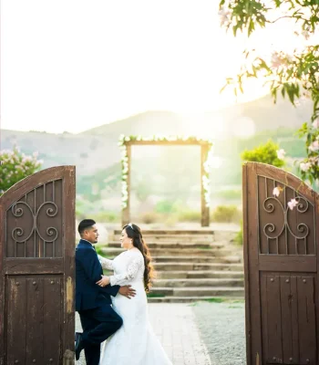Wedding couple at a rustic wooden gate — bride and groom embracing at a wooden garden entrance with string lights beyond