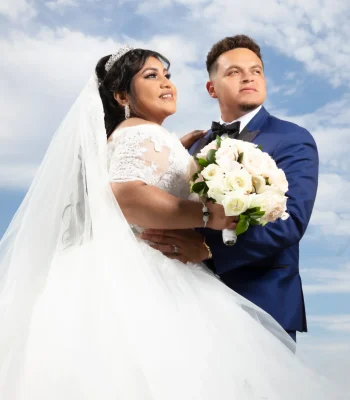Wedding portrait against cloudy sky — groom holding bride, both looking up, white bouquet in foreground