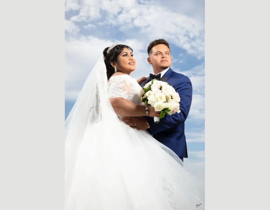 Wedding portrait against cloudy sky — groom holding bride, both looking up, white bouquet in foreground
