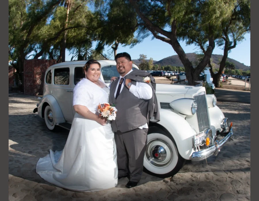 Wedding couple with vintage car (mountain backdrop) — bride and groom posing beside a vintage car with hills in the background