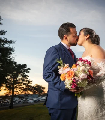 Wedding couple kissing at sunset — groom and bride kissing outdoors with a colorful bouquet, sunset in background