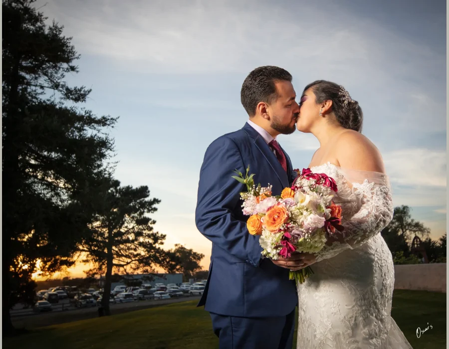 Wedding couple kissing at sunset — groom and bride kissing outdoors with a colorful bouquet, sunset in background