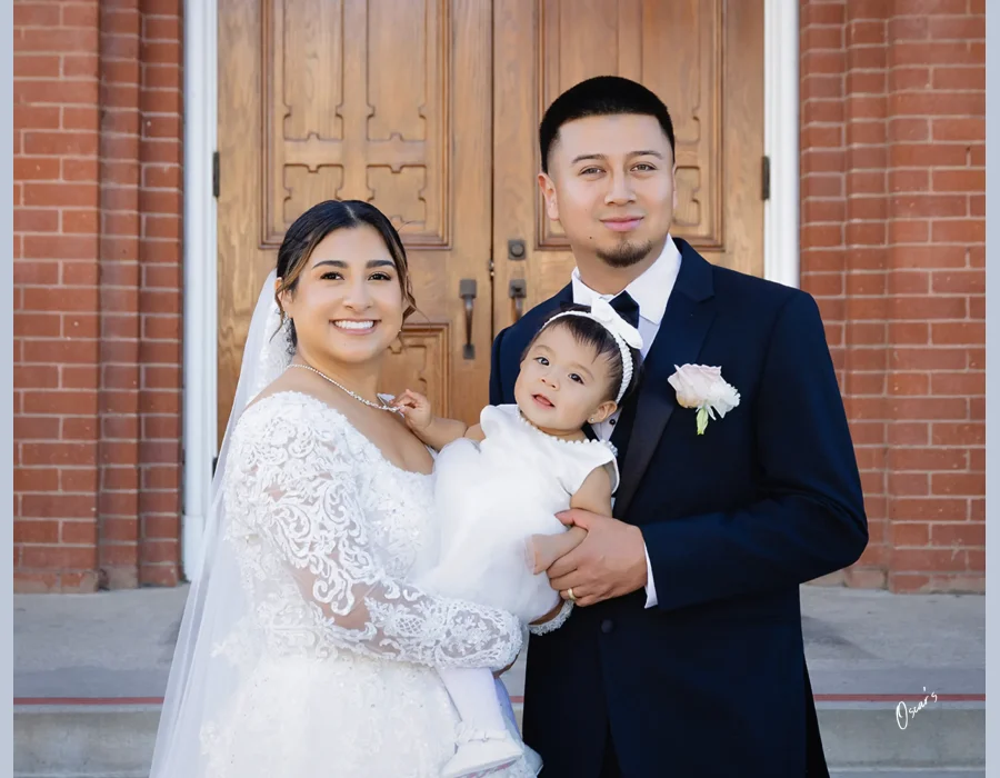 Wedding family portrait at church entrance — bride and groom holding their baby daughter in front of wooden church doors