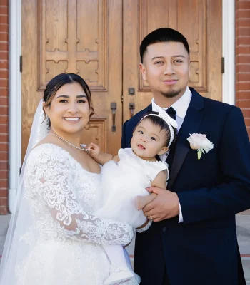 Wedding family portrait at church entrance — bride and groom holding their baby daughter in front of wooden church doors