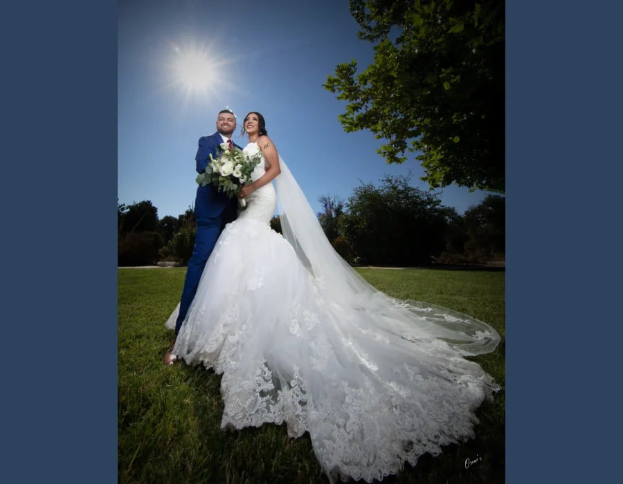 Wedding portrait with dramatic sunburst — bride and groom laughing together outdoors, sun flare and long lace train