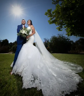 Wedding portrait with dramatic sunburst — bride and groom laughing together outdoors, sun flare and long lace train