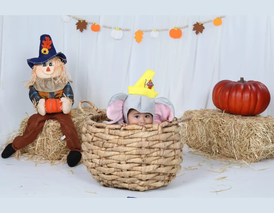 An infant wearing a grey elephant costume with pink ears and a yellow circus hat peeking out from inside a large woven wicker basket, set against an autumn-themed backdrop featuring a scarecrow, hay bales, pumpkins, and a fall leaf banner.