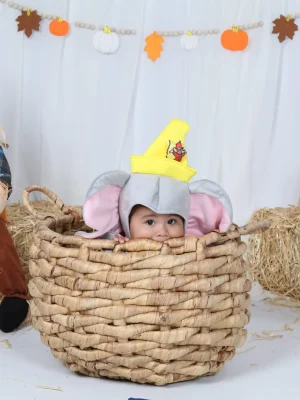 An infant wearing a grey elephant costume with pink ears and a yellow circus hat peeking out from inside a large woven wicker basket, set against an autumn-themed backdrop featuring a scarecrow, hay bales, pumpkins, and a fall leaf banner.