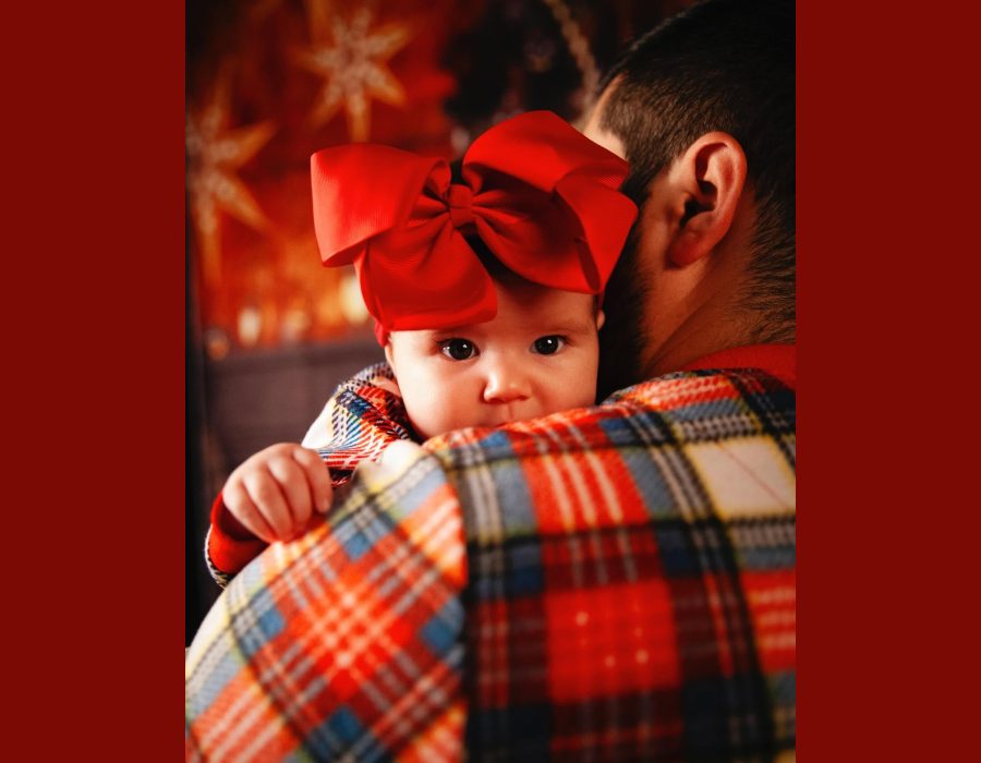 close-up of a father holding an infant wearing a large red bow headband and plaid outfit, warm Christmas-lights backdrop