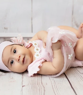 A smiling baby girl wearing a pink floral dress and a matching pink headband while lying on her back on a wooden floor.