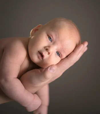 A newborn baby with wide eyes resting comfortably in the palm of an adult's hand against a dark grey studio background.