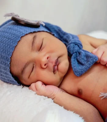 A newborn baby sleeping peacefully on a white textured blanket, wearing a blue knitted cap with a side bow and matching blue pants.