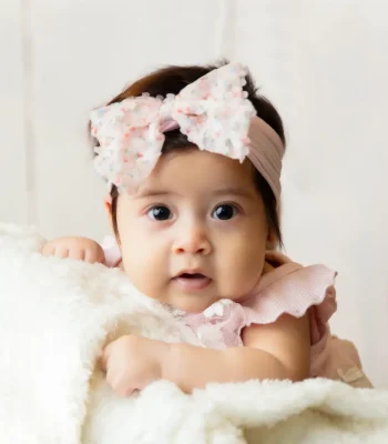 A close-up studio shot of a baby girl with big dark eyes lying on a white fluffy blanket, wearing a pink ruffled dress and a large floral bow headband.