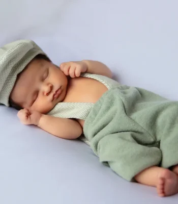 A newborn baby sleeping peacefully on a white blanket, wearing green overalls and a matching newsboy cap.
