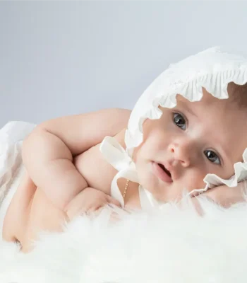 A close-up studio shot of an alert baby lying on a white faux-fur blanket while wearing a white ruffled bonnet and diaper.