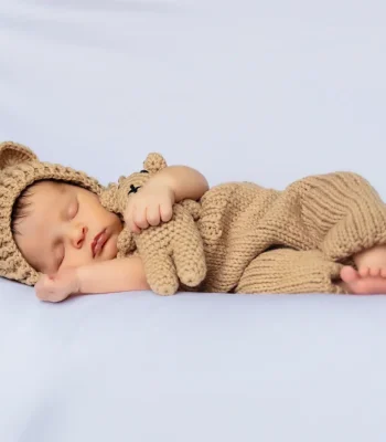 A sleeping newborn baby curled up on a white surface, dressed in a knitted brown bear outfit with matching bear ears and holding a tiny teddy bear.