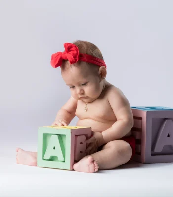 An infant girl wearing a red outfit and a red headband, sitting on the floor and playing with large pastel alphabet blocks spelling "ABC".