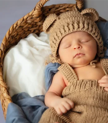 A newborn baby sleeping soundly inside a woven basket lined with a white cloth, blue blanket, and a knitted brown outfit with bear ears.