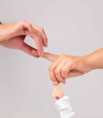 A tender studio shot of adult hands holding and gently grasping the tiny finger of a baby wearing a white sleeve.