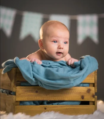 A baby lying on their tummy inside a rustic wooden crate draped with a blue blanket, with pastel bunting banners hanging in the background.