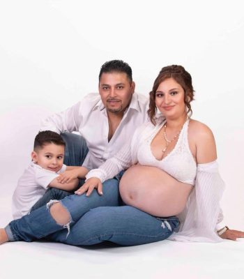 pregnant mother in white lace top, husband, and young son sitting together against a white studio backdrop