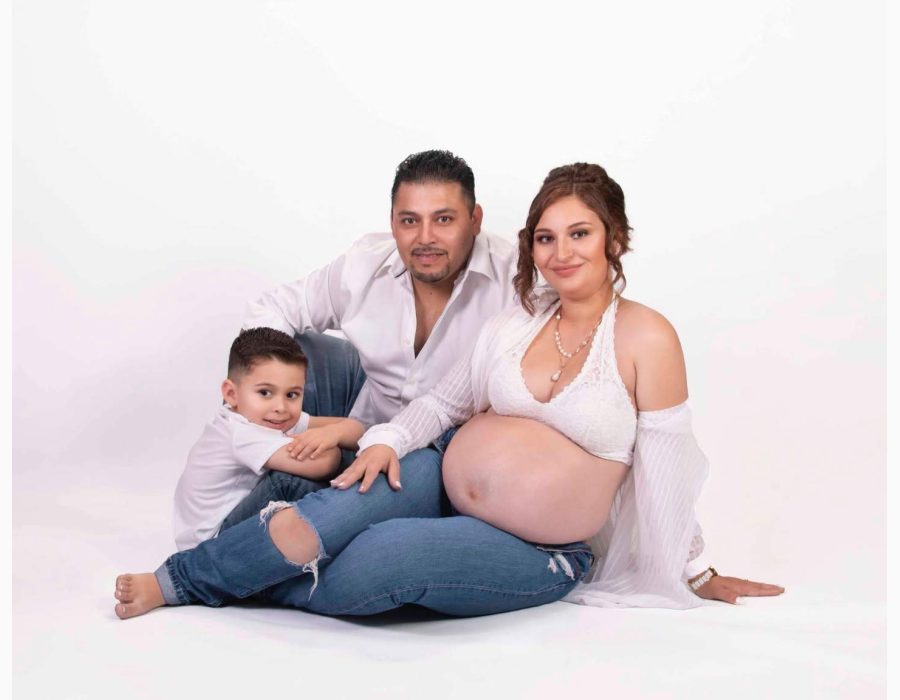 pregnant mother in white lace top, husband, and young son sitting together against a white studio backdrop
