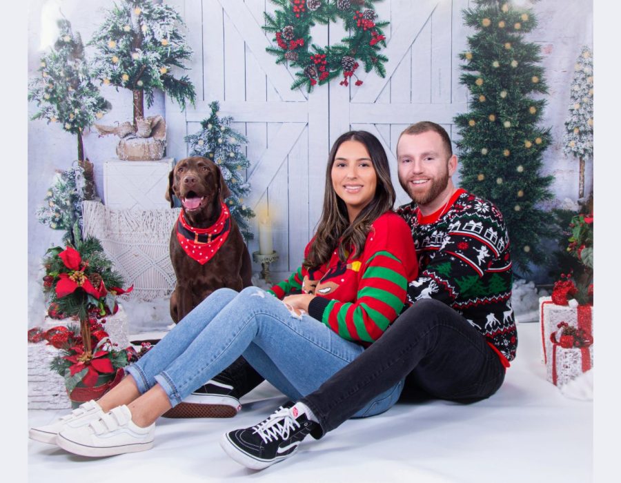 A couple posing with their chocolate Labrador dog in front of a snowy winter holiday backdrop with Christmas trees, a wreath, and presents.