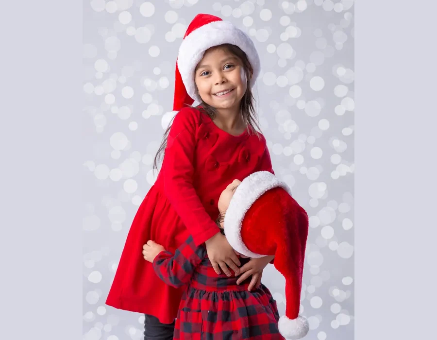 A young girl in a red dress and Santa hat smiling while hugging another child whose face is hidden under a Santa hat, set against a bokeh background.