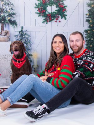 A couple posing with their chocolate Labrador dog in front of a snowy winter holiday backdrop with Christmas trees, a wreath, and presents.