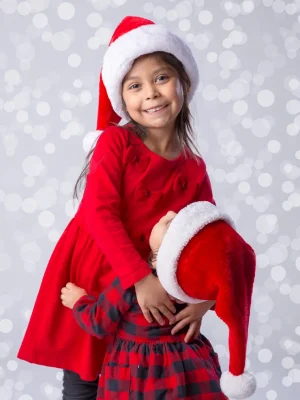 A young girl in a red dress and Santa hat smiling while hugging another child whose face is hidden under a Santa hat, set against a bokeh background.