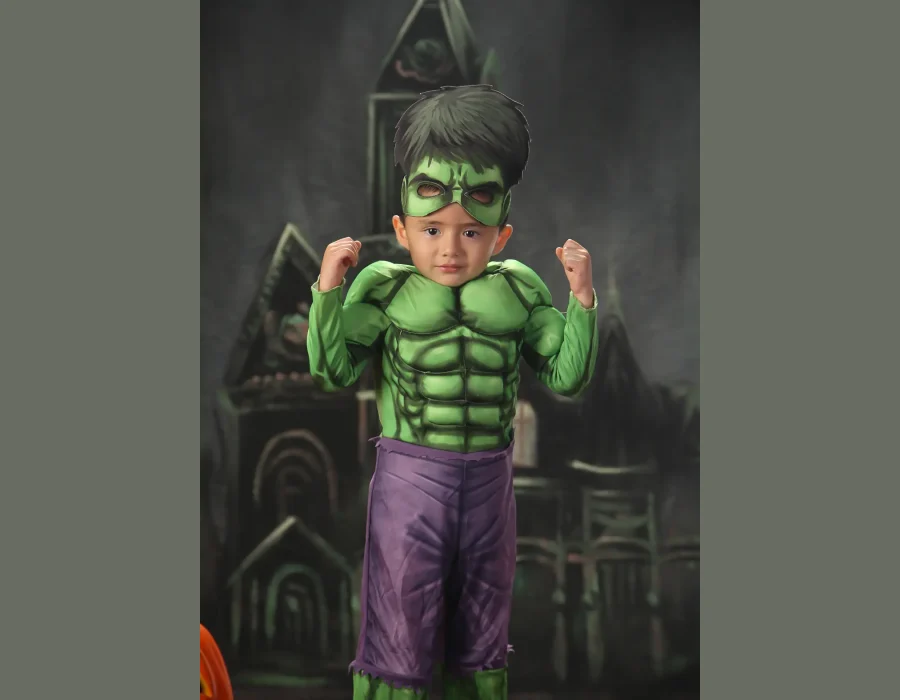 A young boy in a Hulk costume striking a flexed muscle pose with both arms up, standing against a dark castle and pumpkin backdrop.