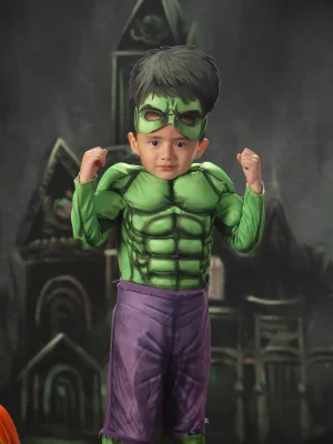 A young boy in a Hulk costume striking a flexed muscle pose with both arms up, standing against a dark castle and pumpkin backdrop.