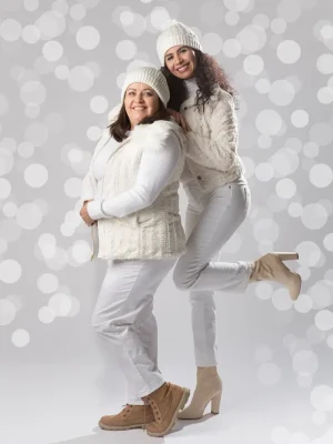 Two women wearing matching white winter outfits, white knit beanies, and boots posing together playfully against a bright bokeh background.