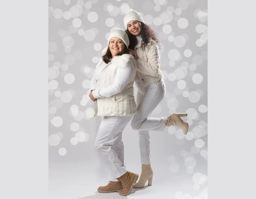 Two women wearing matching white winter outfits, white knit beanies, and boots posing together playfully against a bright bokeh background.