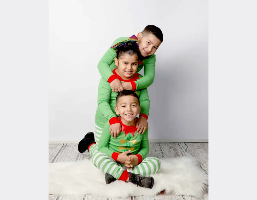 Three children dressed in matching green and red elf pajamas stacked on top of each other, sitting on a white rug next to a frosted miniature Christmas tree and gold wrapped presents.