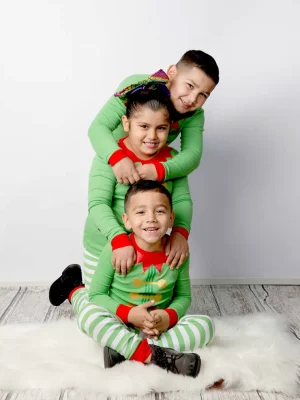 Three children dressed in matching green and red elf pajamas stacked on top of each other, sitting on a white rug next to a frosted miniature Christmas tree and gold wrapped presents.