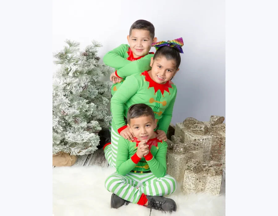 Three children in elf pajamas smiling and stacking together playfully on a white rug with a wooden floor background.