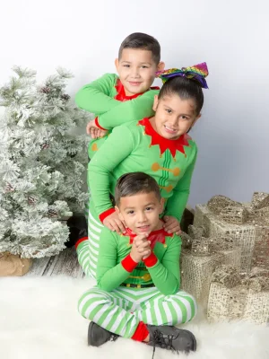 Three children in elf pajamas smiling and stacking together playfully on a white rug with a wooden floor background.