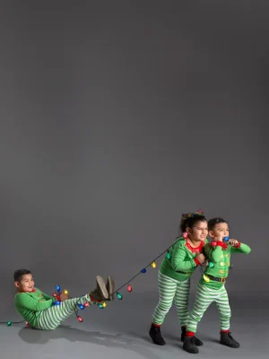 Two children in matching green and red elf pajamas pull on a string of colorful Christmas lights, while a third child in identical pajamas sits on the floor holding the other end and smiling.