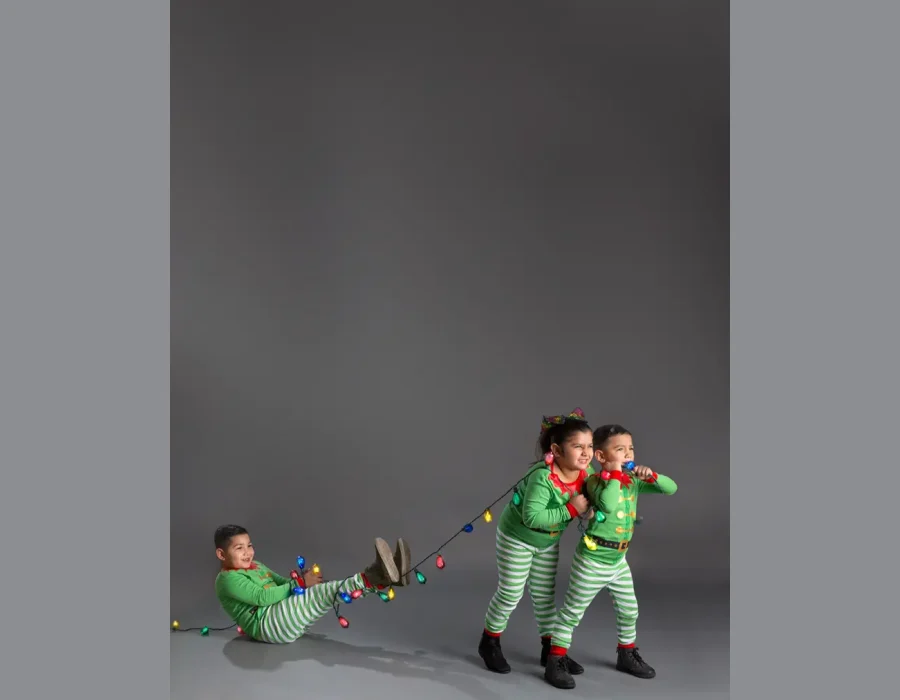 Two children in matching green and red elf pajamas pull on a string of colorful Christmas lights, while a third child in identical pajamas sits on the floor holding the other end and smiling.