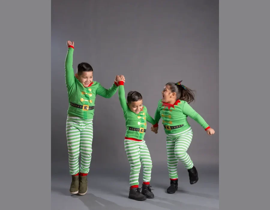 Three children in matching green and red elf pajamas holding hands and jumping playfully in unison against a grey background.
